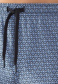 Close-up van blauw gemusterde stof met kleine bloemachtige vormen en een zwarte trekkoord die door een zwart metalen oogje is geregen.