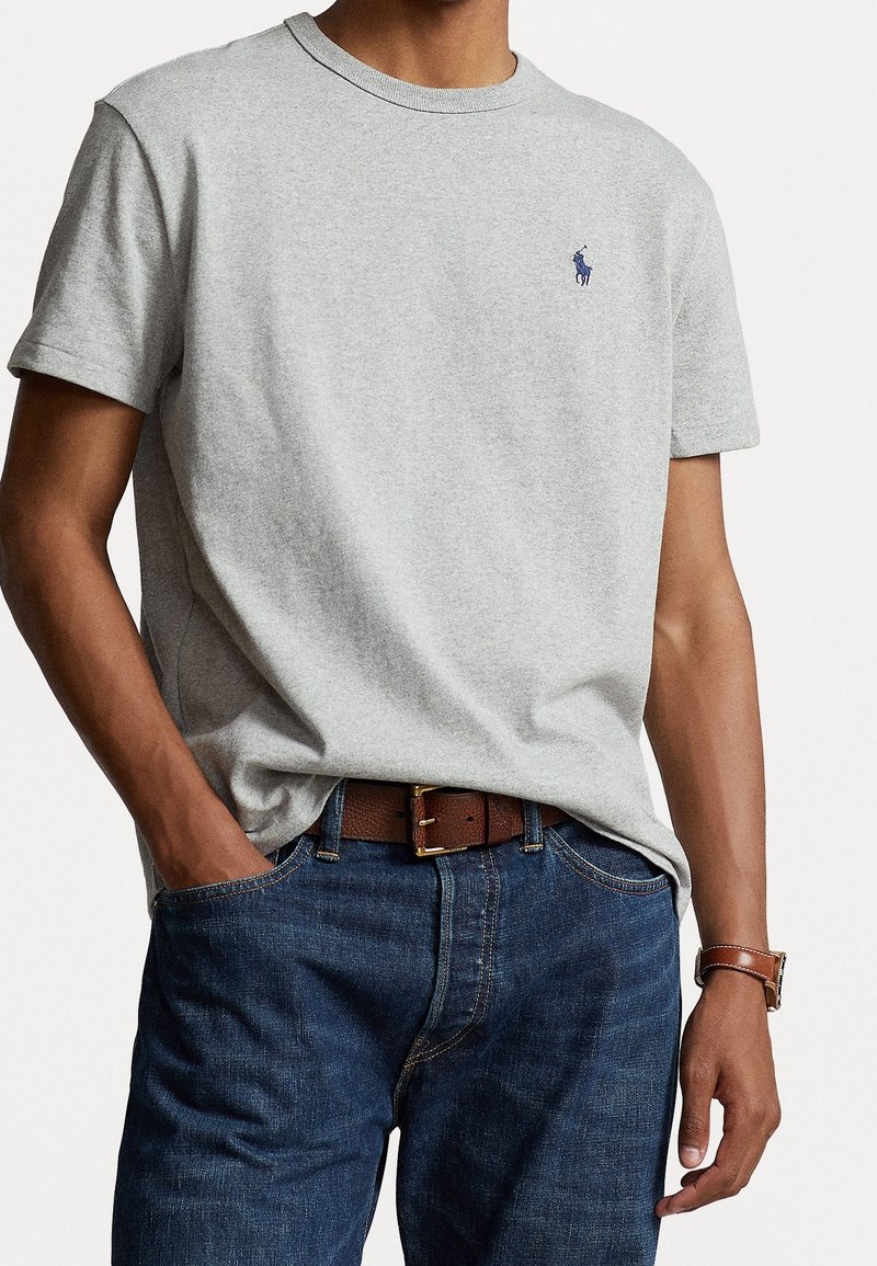 Šedé bavlněné tričko s krátkým rukávem a kulatým výstřihem, na levé hrudi má malé modré logo, ve dvojici s tmavými džíny a hnědým opaskem.