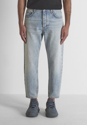Hombre con vaqueros azul claro desgastados y zapatillas gris oscuro, de pie frente a un fondo liso.