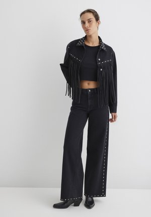 Black denim jacket with fringe and stud accents; paired with high-waisted black pants featuring stud detailing along the side seams.