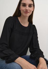 Juoda ilgos rankovės bluzonė su apvaliu iškirpimu, kuriame yra tekstūruotos nėrinių detalės priekyje. Pūkuotos rankovės su susiraukšlėmis.