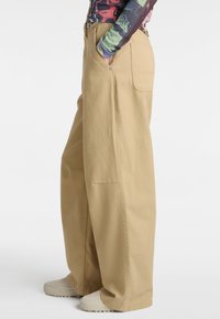 Pantalones de pierna ancha beige con un acabado texturizado, que cuentan con bolsillos laterales y un cierre de botón, combinados con calzado en tonos neutros.