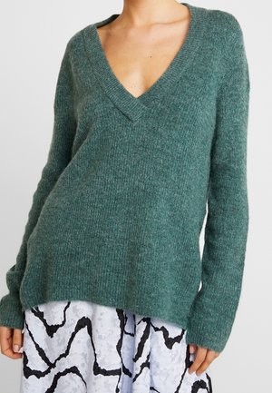 Frau trägt einen tealfarbenen, gerippten V-Ausschnitt-Pullover, kombiniert mit einem weißen Rock mit einem schwarzen abstrakten Muster.