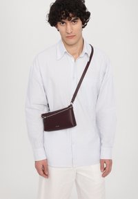 Burgunderrød lær crossbody-veske med glidelås og justerbar stropp, båret over en stripete skjorte med knapper. Enkel og stilren design.
