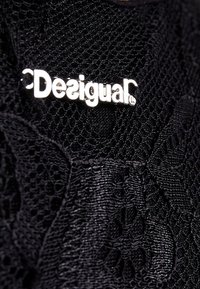 Černá texturovaná tkanina s propracovanými vzory a prominentně zobrazeným kovovým stříbrným logem "Desigual".