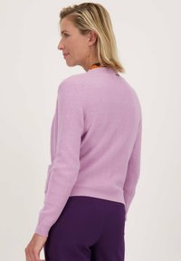 Lila geribde cardigan met een ronde halslijn en lange mouwen. Draagt met paarse broek, die een subtiel label op de achterkant heeft.