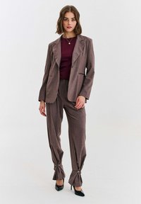 Hnedý, štruktúrovaný oblek s priliehavým sakom s výstrihom s výrezmi a širokými nohavicami so zapínaním na gombíky na leme. Vzhľad dopĺňa bordový top.