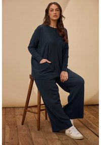 Maglione blu scuro, morbido e ampio, con tasche laterali, abbinato a pantaloni a gamba larga. Modella seduta su uno sgabello di legno, indossando sneakers bianche.