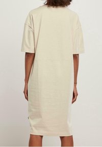 Beige puuvillainen ylisuurikokoinen t-paitamekko, jossa on lyhyet hihat, pyöreä pääntie ja suora leikkaus. Sileä pinta ja näkyvät ompelusaumat.