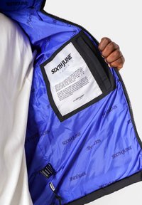 De binnenkant van de ritsjack is gemaakt van zwart en blauw stof, met een logo dat door het hele jack is gedrukt, en bevat een zwart label met verzorgingsinstructies.