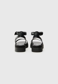 Sandales en cuir noir avec une large bride à la cheville, deux brides croisées à l'avant et une semelle épaisse au toucher daim.