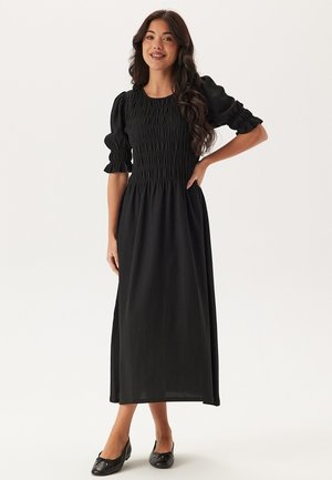 Zwarte maxi-jurk met een gerimpelde lijfje, poffende mouwen en een gestructureerd stoffenpatroon. Gestyled met eenvoudige zwarte flats.