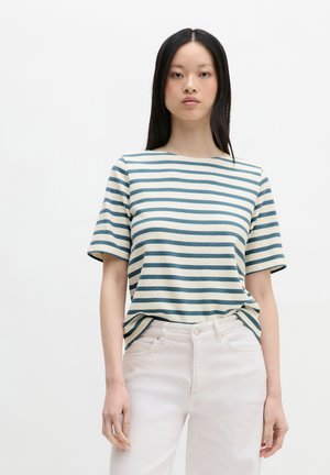 Junge Frau mit langen dunklen Haaren, die ein blau-weiß gestreiftes kurzärmeliges Oberteil und weiße Hose trägt, steht vor einem einfarbigen Hintergrund.