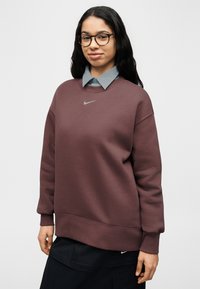 Μπορντό φούτερ με στρογγυλή λαιμόκοψη και μακριά μανίκια, με γκρι λογότυπο Nike στο στήθος. Φορεμένο πάνω από ένα ανοιχτού μπλε πουκάμισο με γιακά.