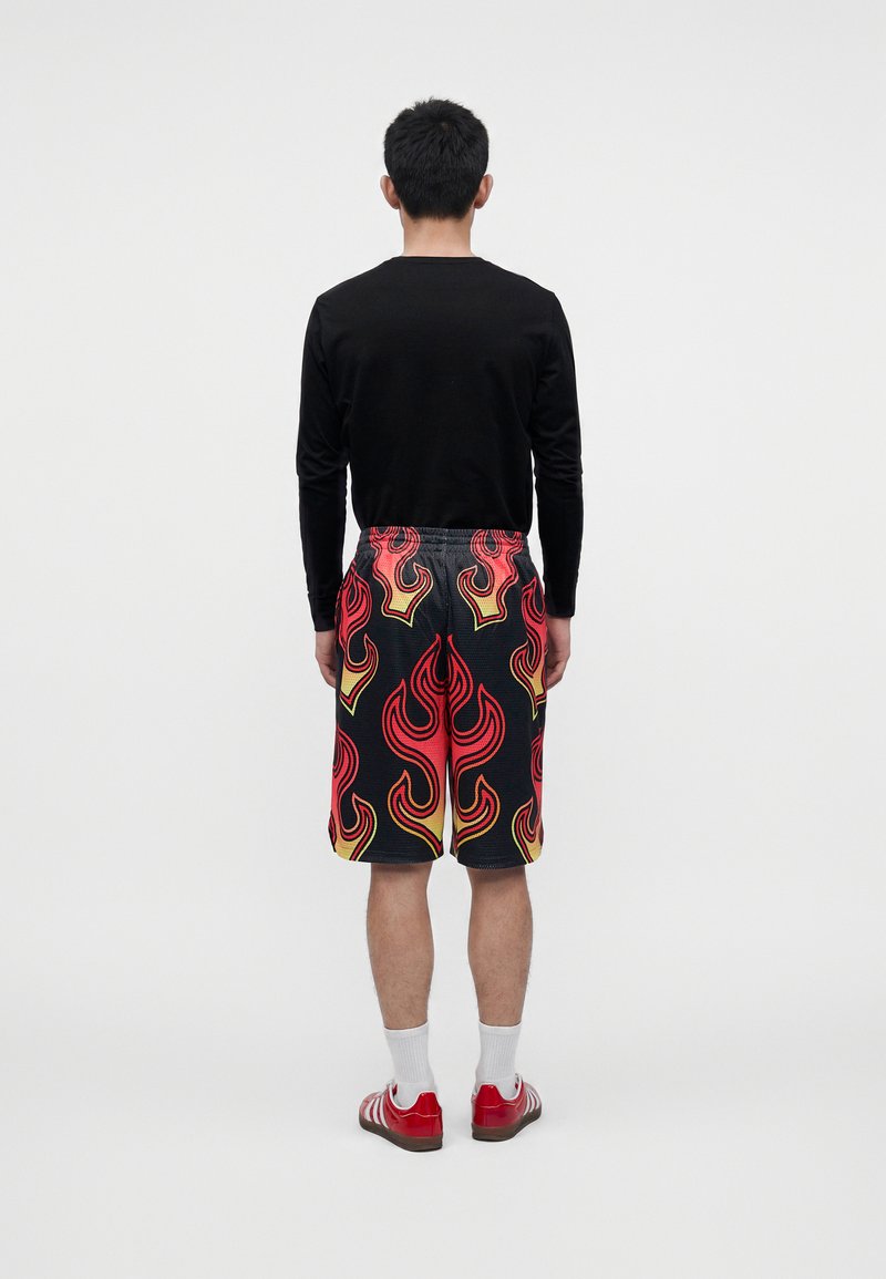 Uomo di spalle che indossa una maglia nera a maniche lunghe, pantaloncini neri con motivo di fiamme rosse e gialle, calzini bianchi e sneakers rosse.