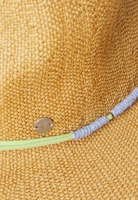 Sombrero de paja en color tostado natural con una textura tejida. Presenta una banda decorativa en verde y lavanda, además de un acento con un logo plateado.