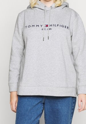 Osoba w jasnoszarej bluzie z kapturem Tommy Hilfiger z troczkami i niebieskich dżinsach, widoczna tułów, bez szczegółów tła.