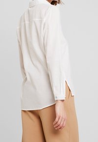 Weißes Langarmhemd mit Kragen, seitlichen Schlitzen und geknöpften Manschetten, aus einem leichten Stoff gefertigt. Glatte Textur und lockere Passform.