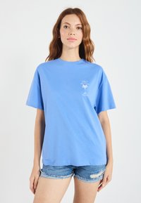 Blue cotton t-shirt with short sleeves, crew neck, and printed palm tree design on the chest. Worn with denim shorts, minimal texture.