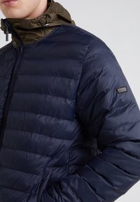 Navy puffer jacket with a quilted design and a detachable olive green hood. Features a front zipper and logo patch on the sleeve.