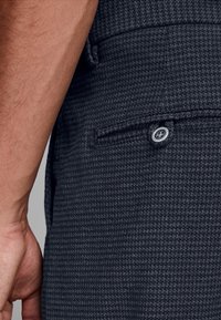 Mörkgrå byxor med ett texturerat hundtandsmönster. Har en sidoficka med en knappstängning. Materialet verkar vara vävt.