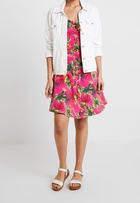 Robe imprimée florale en rose vif, avec des feuilles vertes, un col à volants et un ourlet évasé. Associée à une veste en denim blanche et des sandales.