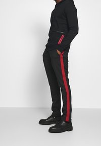 Haut à zip noir avec poche latérale et accents rouges ; pantalon noir assorti avec une rayure rouge sur le côté ; chaussures noires.