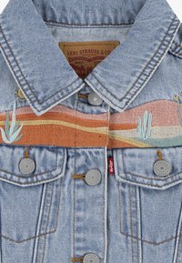 Jeansjacka med ljusblå tvätt, med en färgglad broderad design av berg och kaktusar över bröstet, silverknappar.