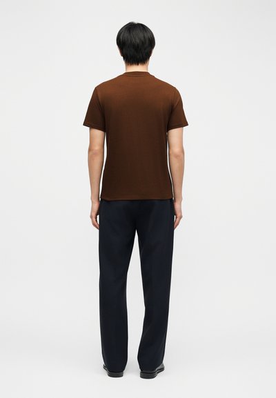 Homme debout de dos portant une chemise marron à manches courtes, un pantalon noir et des chaussures noires sur un fond blanc uni.