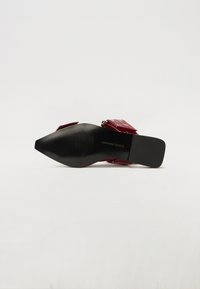 Mule en synthétique rouge avec un nez pointu, semelle texturée noire et une large sangle dotée de quincaillerie métallique et de découpes.