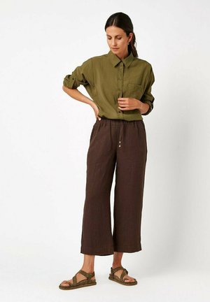 Woman wearing an olive green button-up shirt with rolled sleeves, loose brown cropped pants, and olive green sandals, standing on a white background.