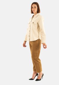 Camisa chaqueta de pana color beige con dos bolsillos frontales y botones marrones, combinada con pantalones de pana marrones y zapatos de tacón negros.