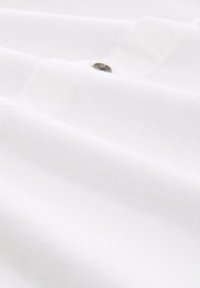 Bijela pamučna tkanina s glatkom teksturom, koja sadrži detalje s gumbom, prikazana u naborima kako bi istaknula svoj pad i laganu kvalitetu.