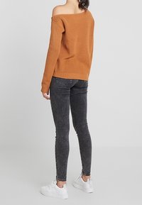 Ein terrakottafarbener, schulterfreier Pullover mit geribbelter Textur, kombiniert mit schwarzen Skinny-Jeans und weißen Sneakern, von hinten betrachtet.