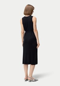 Svart ärmlös midiklänning med figursydd midja, slits vid fållen och mjuk, slät textur. Modell bär silvriga klackskor.