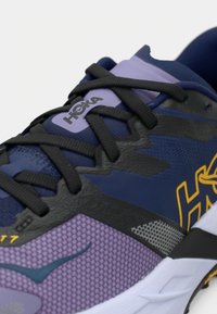 Κοντινή εικόνα ενός μοβ και μπλε παπουτσιού τρεξίματος Hoka με μαύρα κορδόνια και κίτρινο λογότυπο στο πλάι, που δείχνει υλικά με υφή και διχτυωτή επιφάνεια.