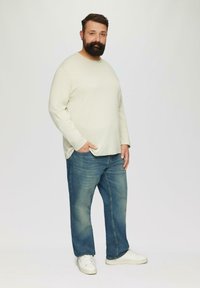 Helles graues Langarmhemd, blaue Jeans und weiße Sneakers. Die gerade geschnittenen Jeans haben subtile Ausbleichungen; das Hemd ist glatt mit einer lässigen Passform.