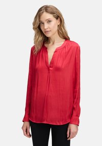 Blouse en soie rouge avec un décolleté en V, des manches longues et un subtil détail de plis aux épaules. Texture lisse et coupe décontractée.