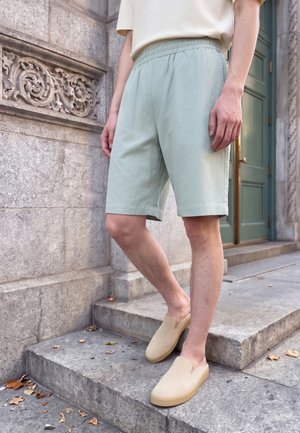 Personne debout sur des marches en pierre, portant un short vert clair, des chaussures beiges sans lacets et une chemise blanche à manches courtes près d’un mur en pierre décoratif et d’une porte verte.