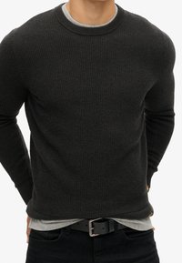 Suéter de punto acanalado gris oscuro con cuello redondo, llevado sobre una camisa gris clara, complementado con jeans negros y un cinturón negro.
