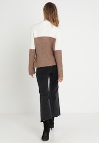 Gebreide trui met een witte bovenkant en een bruine onderkant, met een geribbelde textuur en lange mouwen. Gekleed met zwarte flared broek.