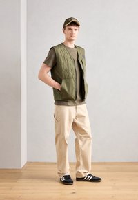 Olive green quilted vest with zip pockets, layered over a brown t-shirt, paired with cream cargo pants and black sneakers featuring white stripes.