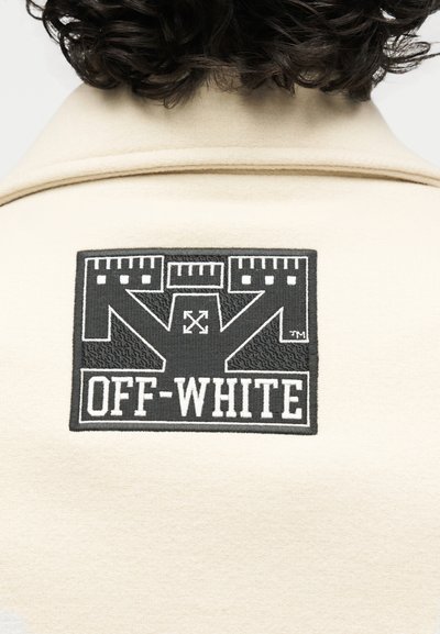 Μπεζ μπουφάν πίσω με μαύρο και λευκό έμπλημα της μάρκας Off-White που δείχνει διασταυρωμένα βέλη και το κείμενο "OFF-WHITE" από κάτω. Ορατά σγουρά σκούρα μαλλιά.