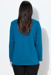 Türkises Langarmshirt mit rundem Halsausschnitt, geradem Schnitt und glatter Textur; getragen mit schwarzen Hosen, von hinten gesehen.