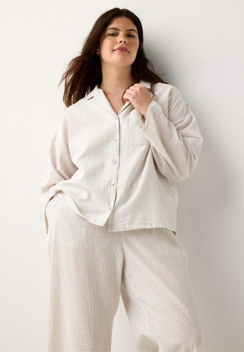 Randig pyjamaset i ljusbeige; lös passform med knapp-up skjorta och ficka, matchande vida byxor. Mjuk textur, avslappnad design.