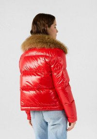 Giacca imbottita rossa con finitura lucida, caratterizzata da un colletto in pelliccia marrone. Il modello è imbottito con cuciture orizzontali e maniche lunghe.