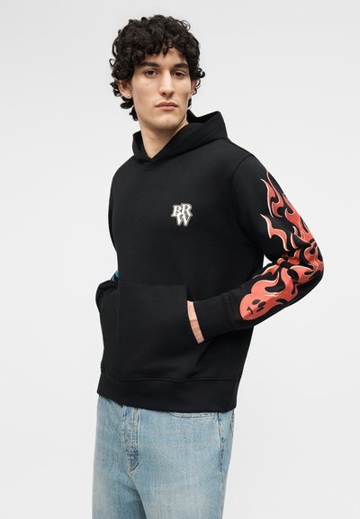 Tânăr purtând un hanorac negru cu grafice de flăcări roșii pe mâneca dreaptă și logo alb „BRW” pe piept, asortat cu blugi albaștri deschiși.