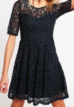Frau trägt ein knielanges schwarzes Spitzenkleid mit Blumenmuster und kurzen Ärmeln, steht mit leicht erhobenem Arm.