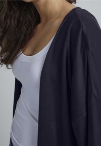 Marinblå stickad kofta med lös passform, öppen front och breda ärmar, lager på en vit tajt linne. Mjuk textur, minimalistisk design.