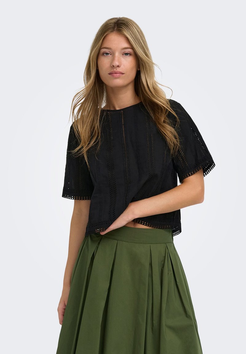 Jeune femme aux longs cheveux blonds portant un haut noir à manches courtes à motifs et une jupe plissée taille haute de couleur vert olive.